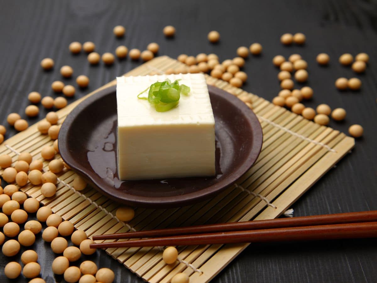 A plate of tofu on a woven mat with chopsticks surrounded by soybeans