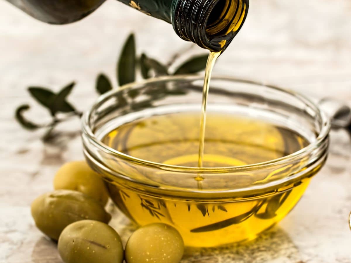 Olive oil being poured into a glass bowl surrounded by fresh olives.