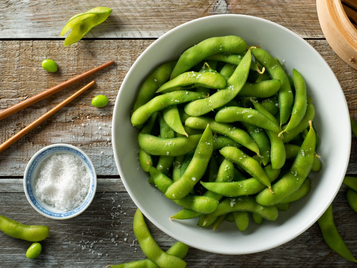 White bowl of steamed edamame on a wooden table with a bowl of salt and a set of chopsticks