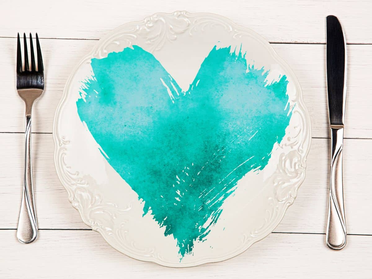 A white plate with a turquoise heart painted on it, flanked by a fork on the left and a knife on the right, on a wooden surface.
