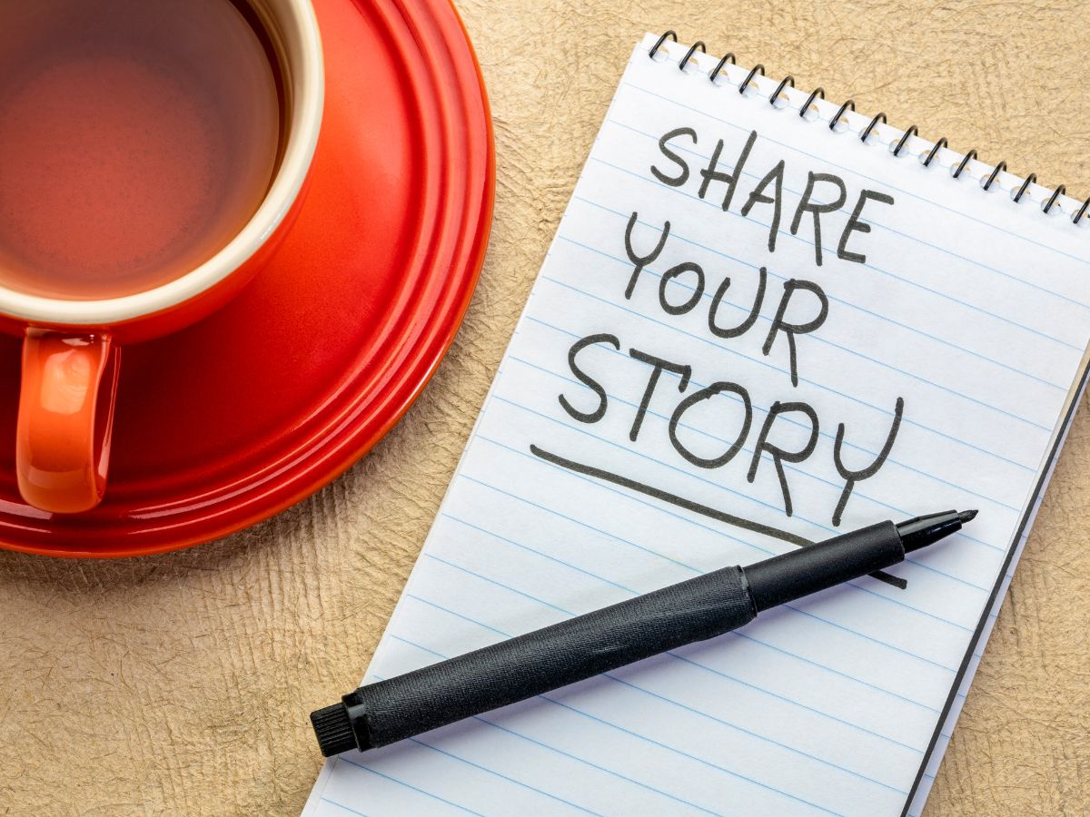"Share Your Story" written on a note pad sitting next to a coffee cup