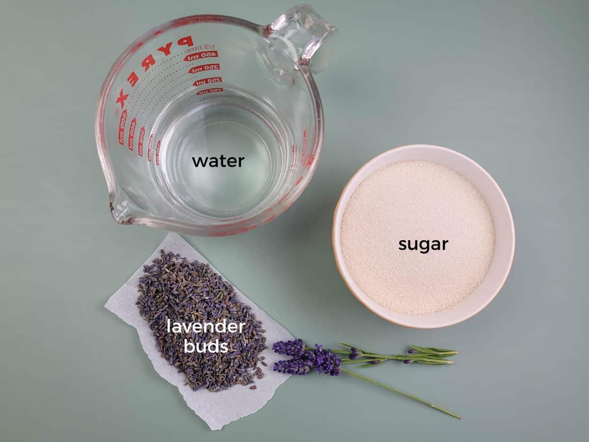 A glass measuring cup of water, a bowl of sugar, and fresh lavender buds to make homemade lavender syrup