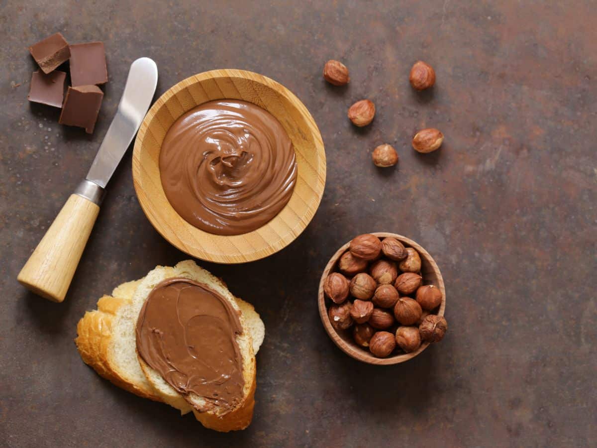 A table with a bowl of chocolate hazelnut spread and bread, surrounded by hazelnuts.