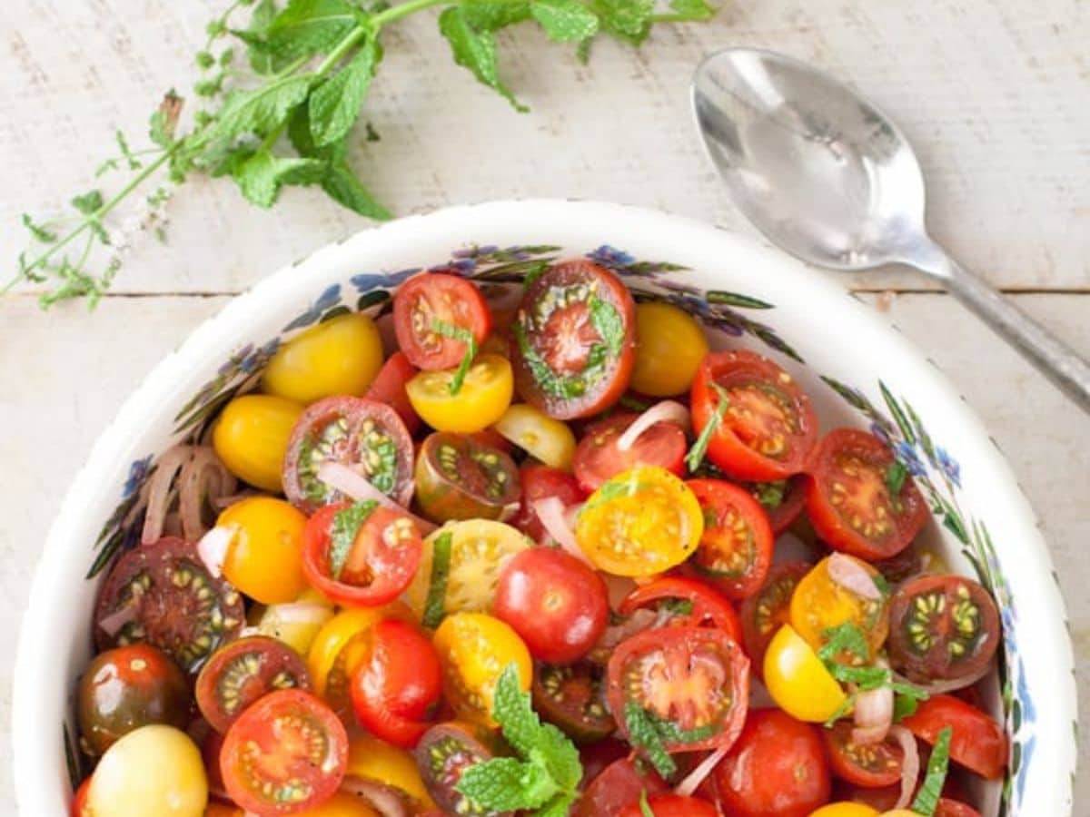 A white bowl of fresh tomato salad with a silver spoon and sprig of fresh mint.