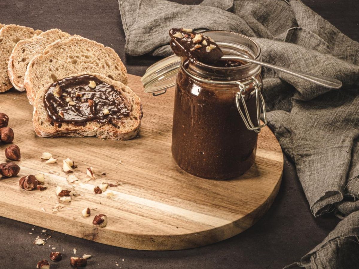 A jar of homemade chocolate spread in a glass jar on a wooden cutting board with nuts.