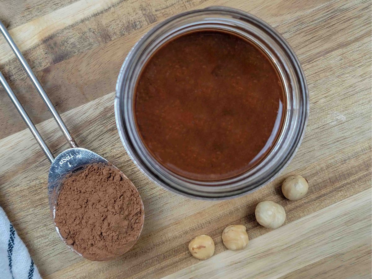 A jar of chocolate hazelnut spread with a spoon of cocoa powder and hazelnuts next to it.