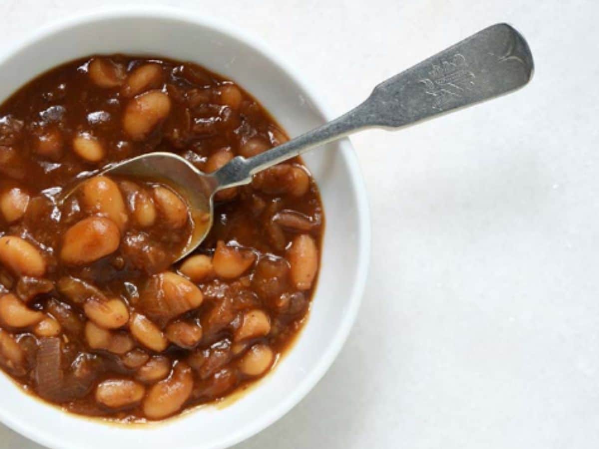 A white bowl of vegan baked beans ready to eat with a silver spoon.
