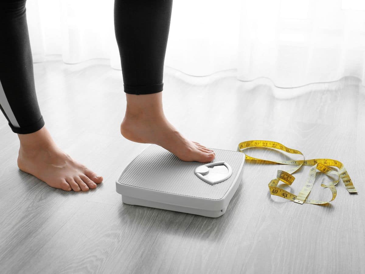 A woman measures her weight on a scale while holding a tape measure, focusing on fitness and health benefits.
