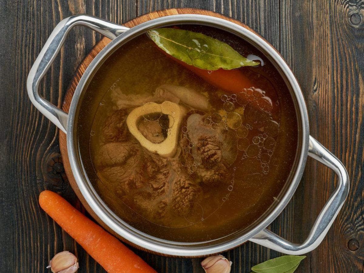 A pot of beef bone broth on a wooden table surrounded by carrots and garlic.