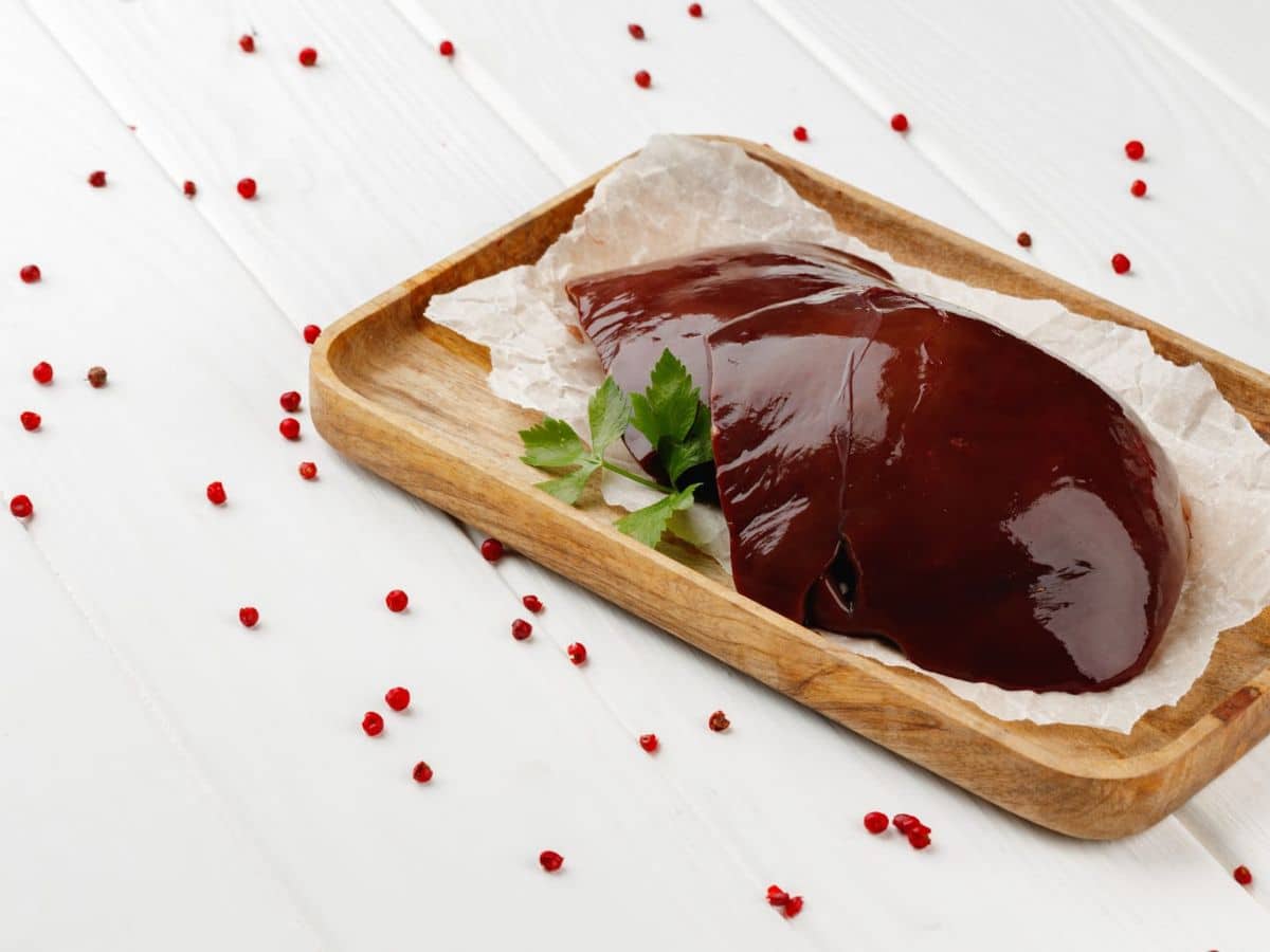 A piece of beef liver on a wooden tray surrounded by red peppercorns.