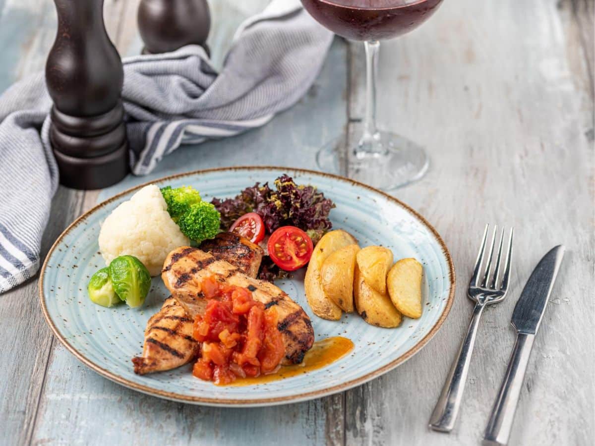 A plate with grilled chicken, potatoes, and tomatoes with a glass of wine nearby.