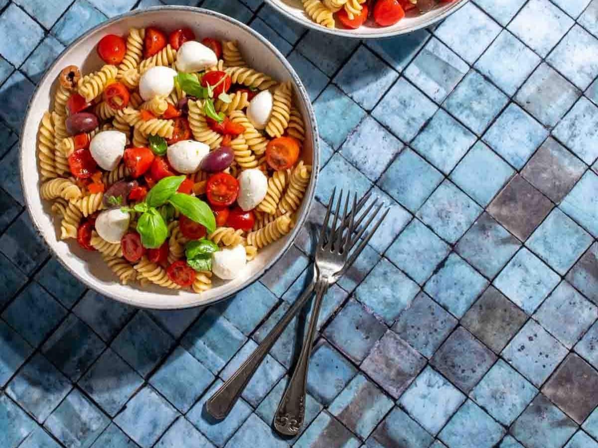 Two bowls of pasta with tomatoes, mozzarella and basil on a tiled background.