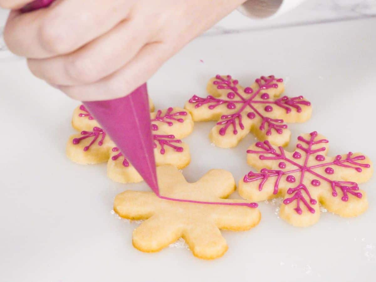 A person adding pink frosting to a snowflake shaped vegan sugar cookie.