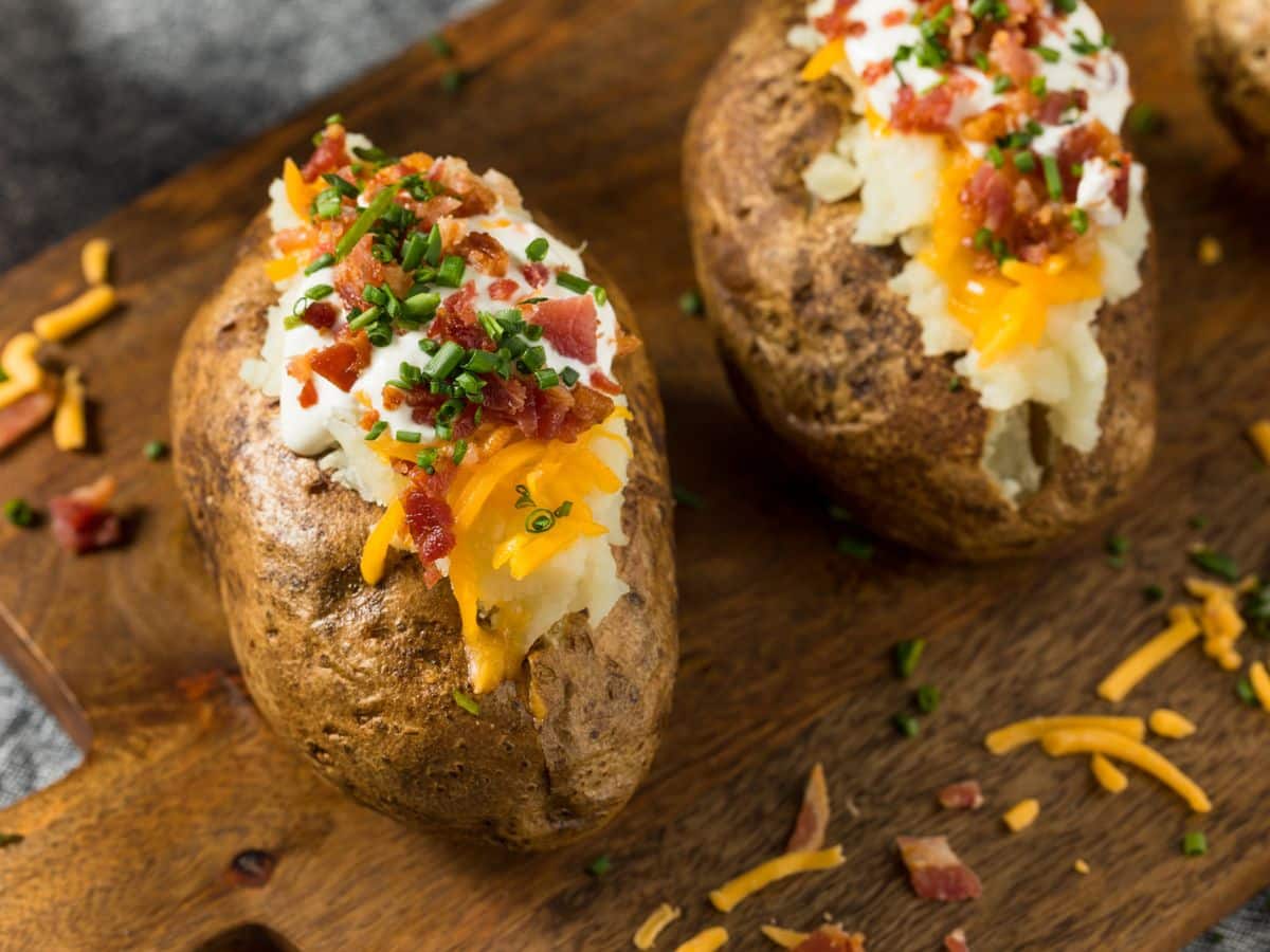 Two baked potatoes with cheese and bacon on a cutting board.