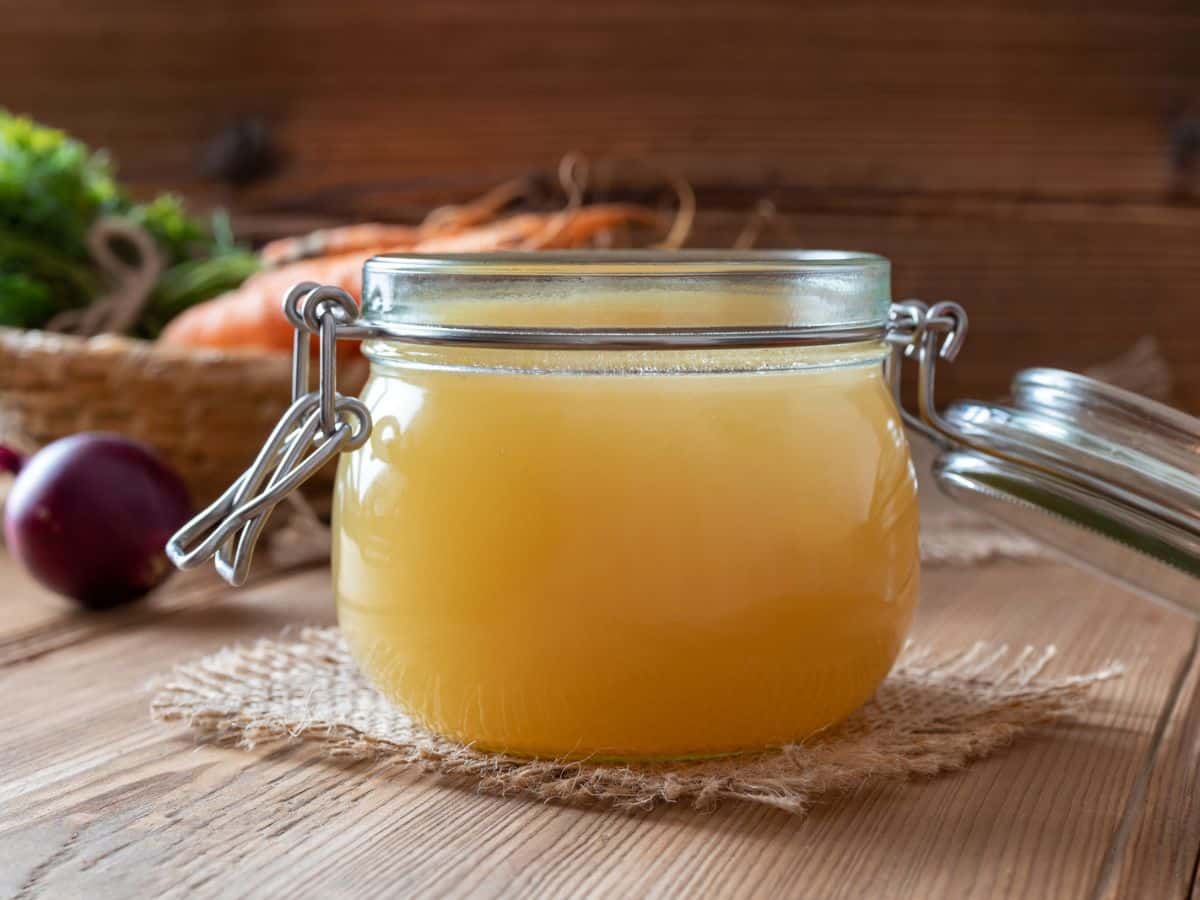 A glass jar filled with homemade chicken stock on a wooden counter with carrots and other vegetables surrounding it.