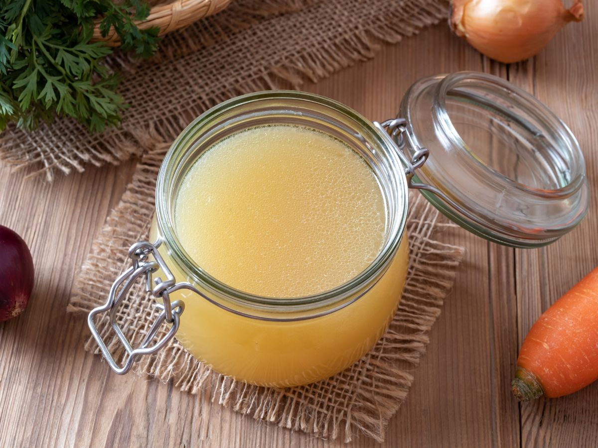 A glass jar of homemade chicken stock, with carrots and onions, on a wooden table.