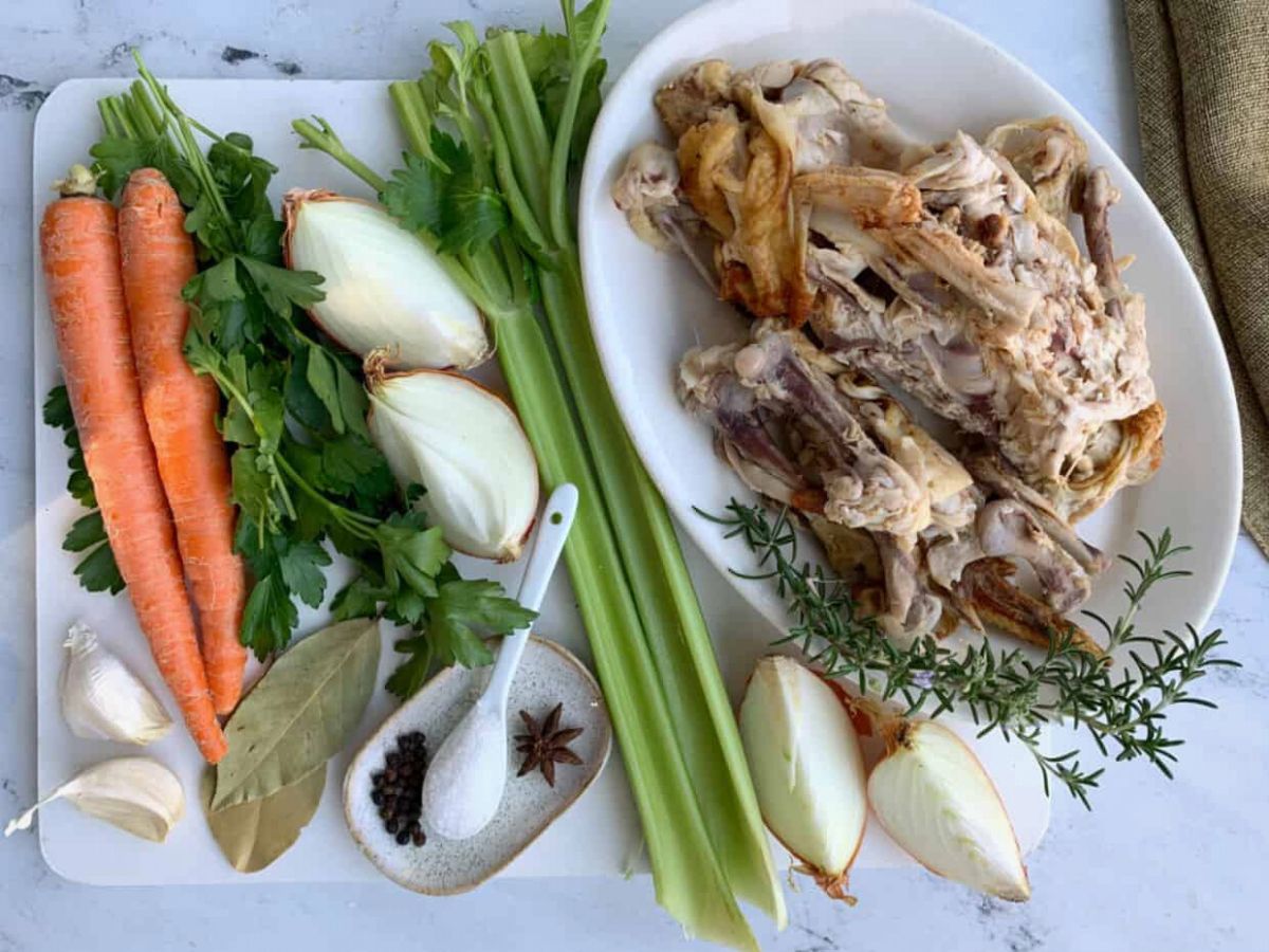 A white plate with carrots, celery and herbs next to a chicken carcass ready to make homemade chicken stock.