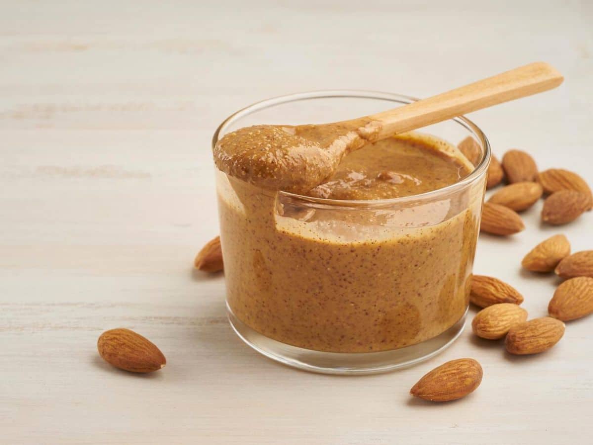 A glass of almond butter surrounded by almonds on a table.