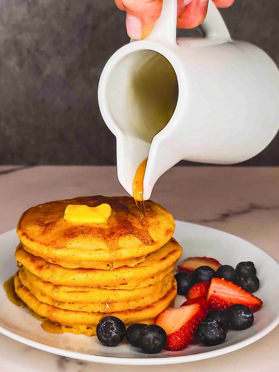 Pouring maple syrup over a stack of dairy-free protein pancakes with butter and berries on the side.