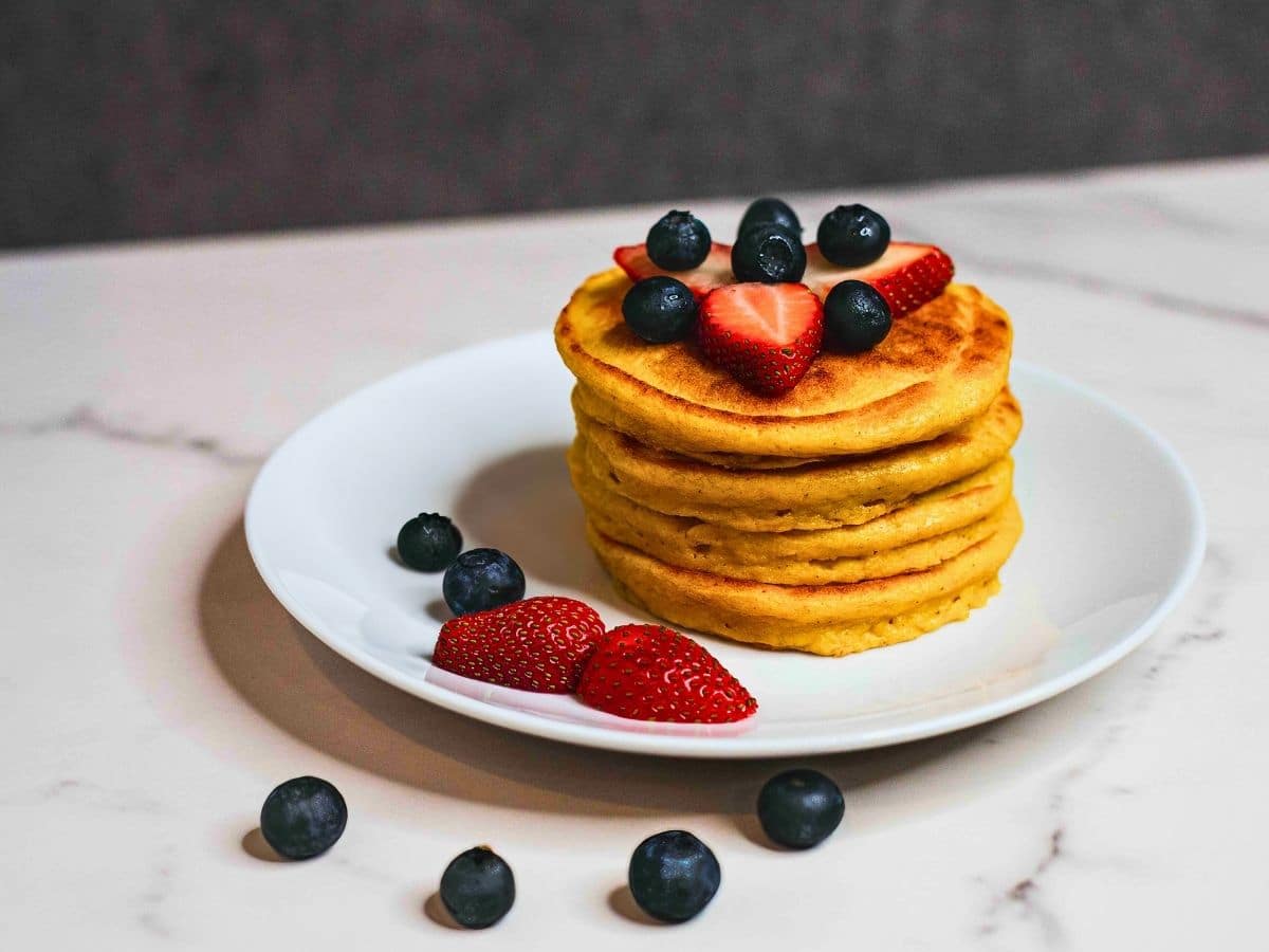 A stack of tofu protein pancakes topped with fresh strawberries and blueberries on a white plate.