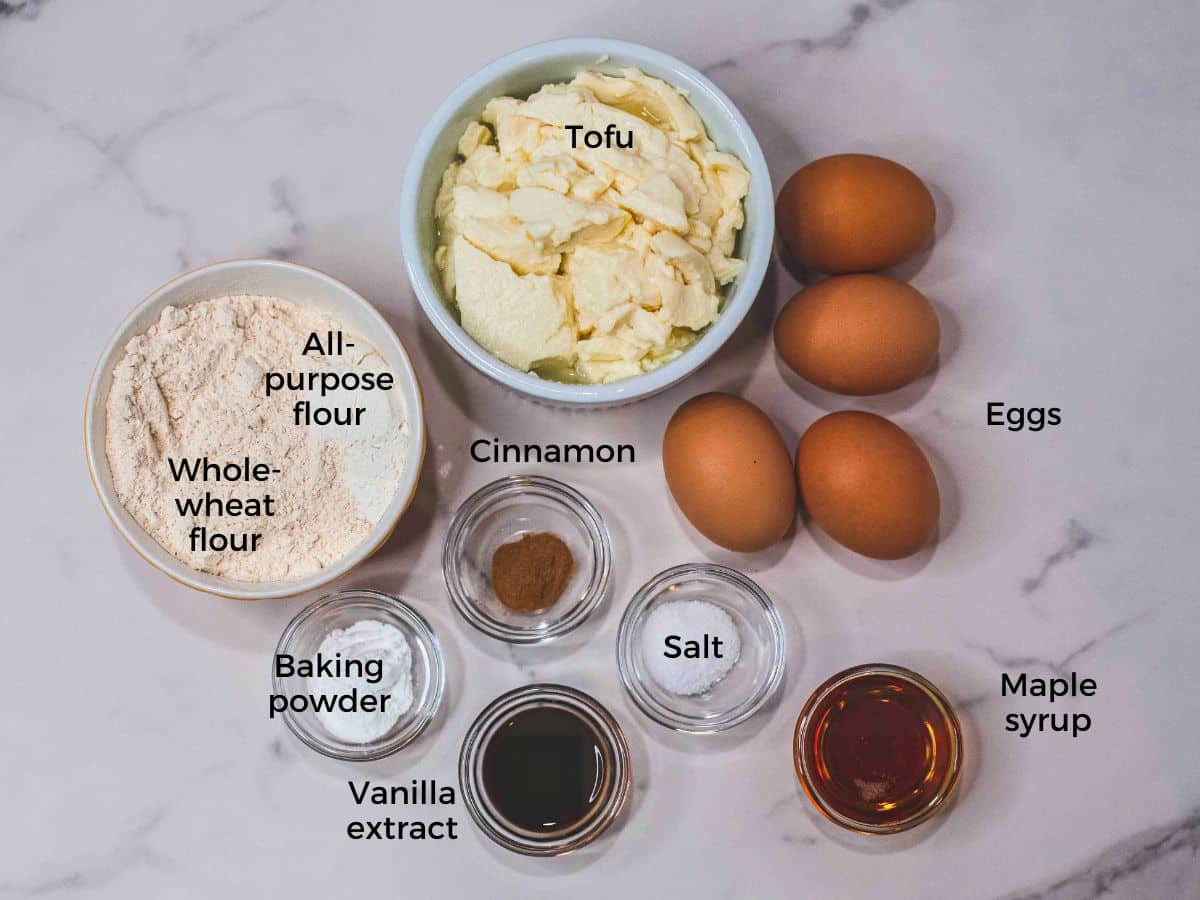 Ingredients for a tofu pancakes recipe laid out on a marble countertop, including tofu, eggs, flours, and flavorings.