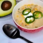 A bowl of white chicken chili garnished with jalapenos and sour cream, accompanied by a halved avocado and a black spoon.