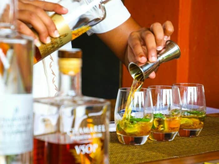 A bartender pours liquor into glasses with ice and mint leaves on a bar counter, with bottles visible in the background.