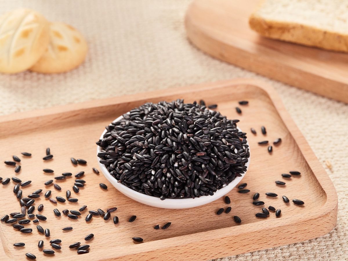 A small white bowl filled with black rice on a wooden tray, with scattered grains around it and slices of bread in the background.