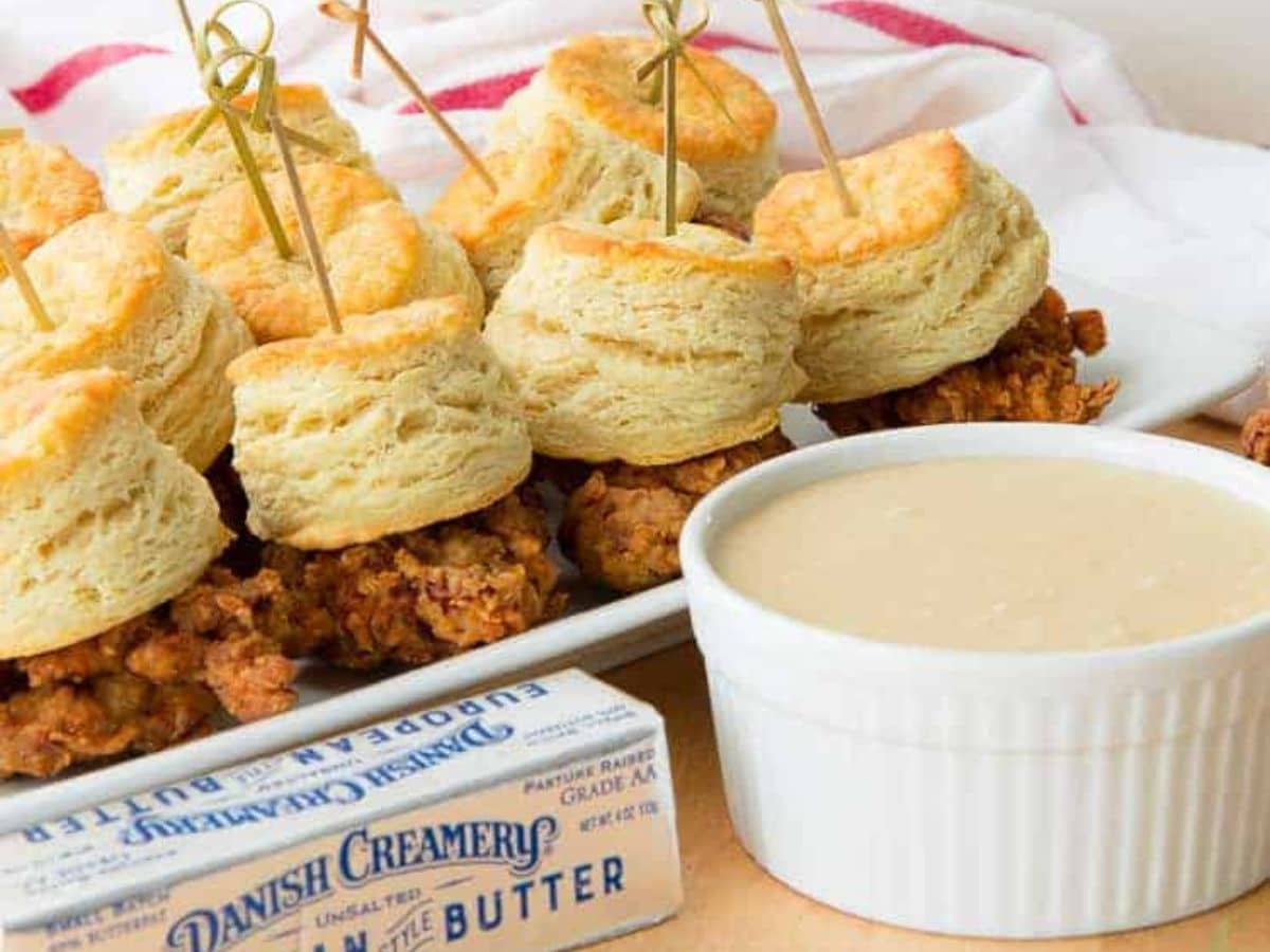 Fried chicken sliders with biscuits on a plate, served with a side of sauce and a block of butter.