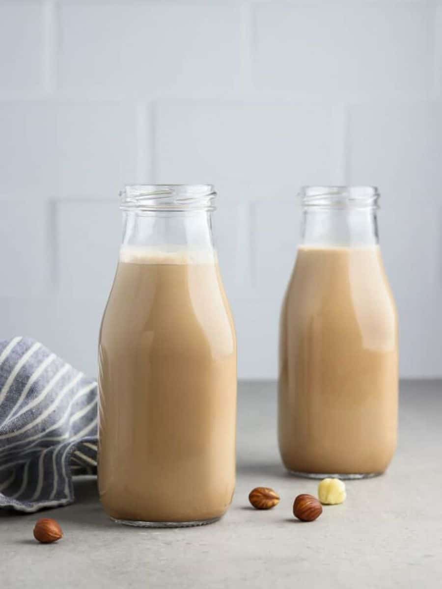 Two bottles of hazelnut milk on a kitchen counter with scattered hazelnuts and a striped cloth beside them.