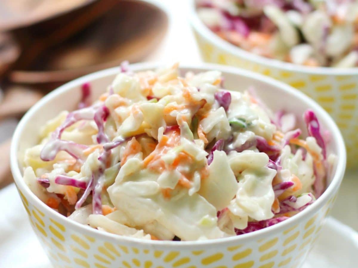Two bowls of creamy coleslaw with shredded red and white cabbage and carrots, served on a wooden table.