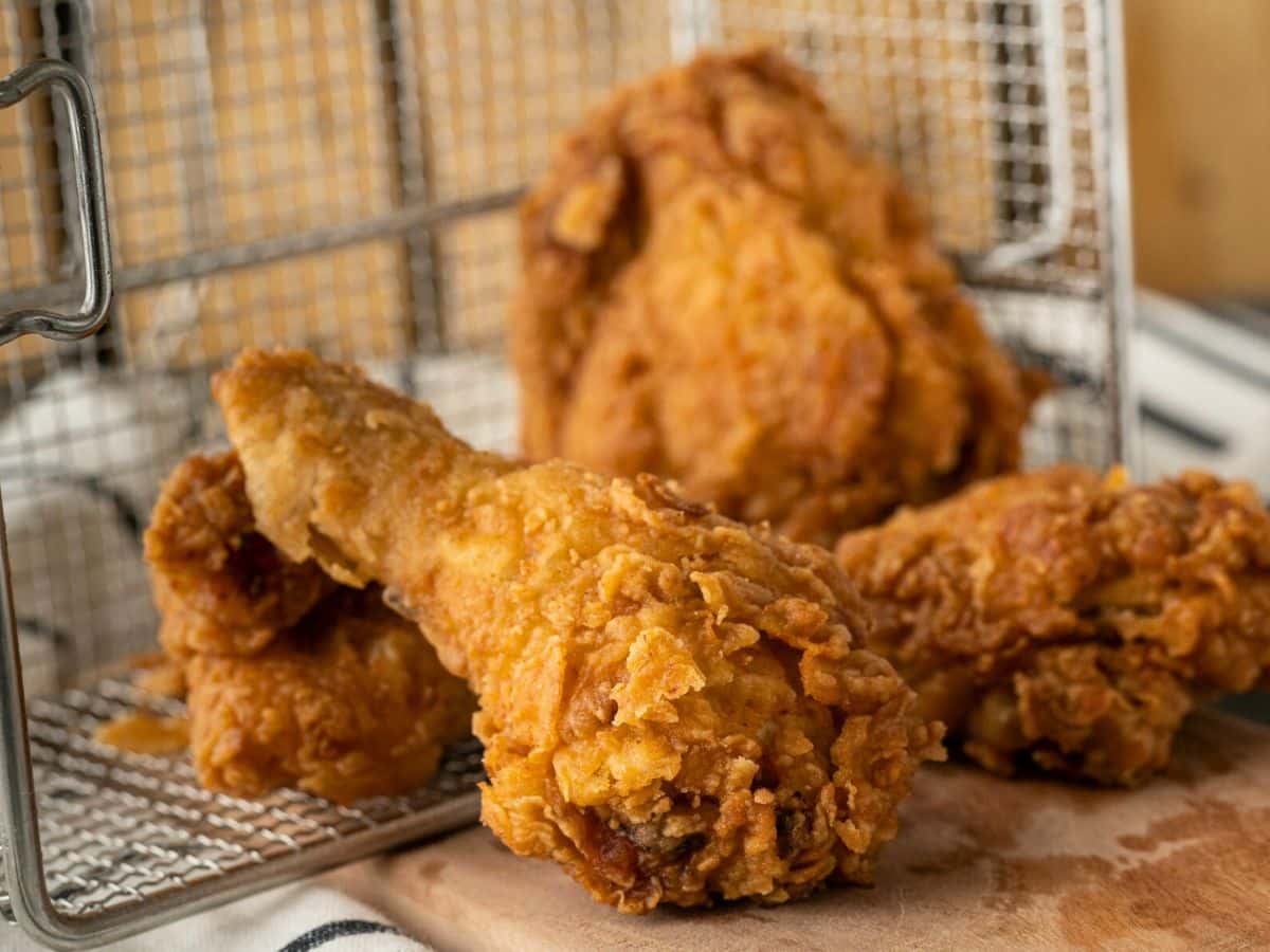 Close-up of crispy fried chicken pieces on a wooden board with a metal basket in the background.