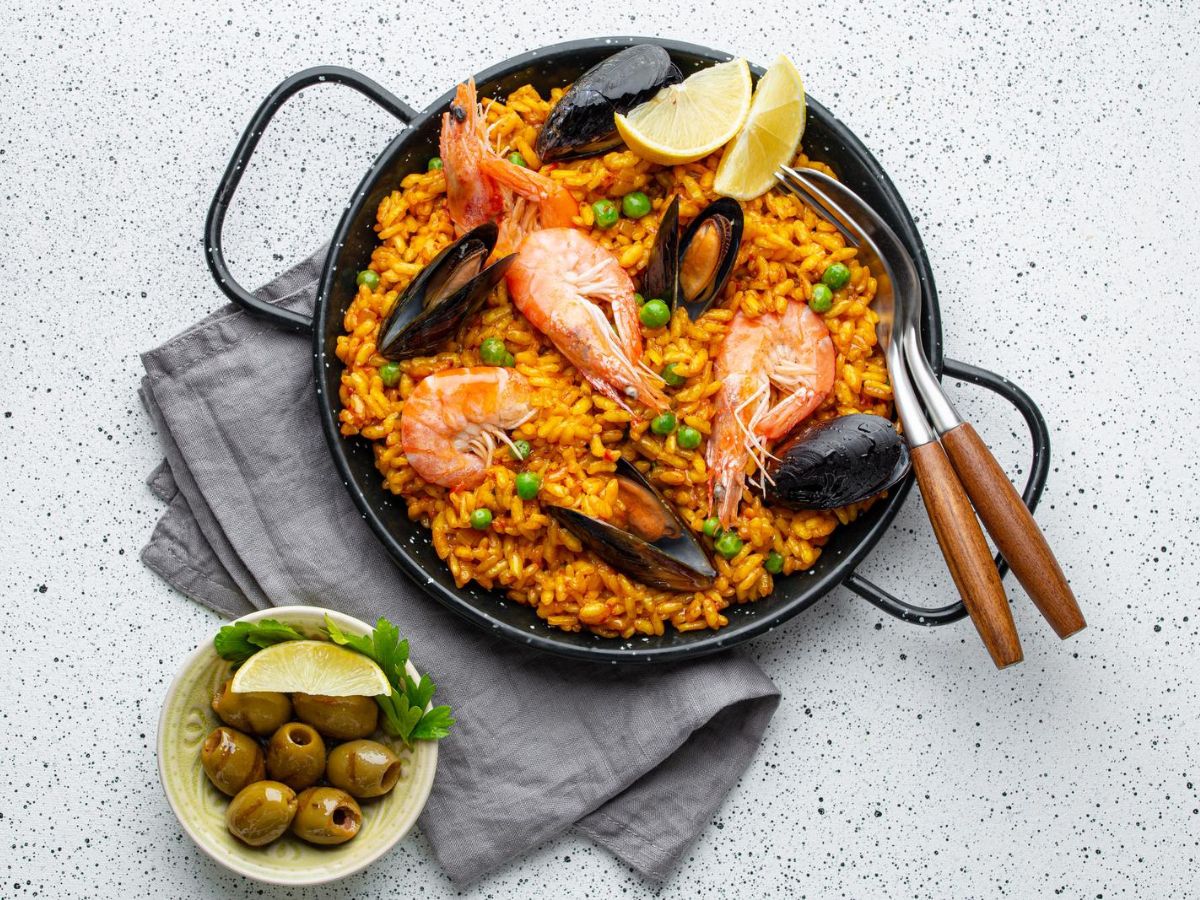 A pan of seafood paella garnished with lemon wedges and served with a side of olives on a speckled table.