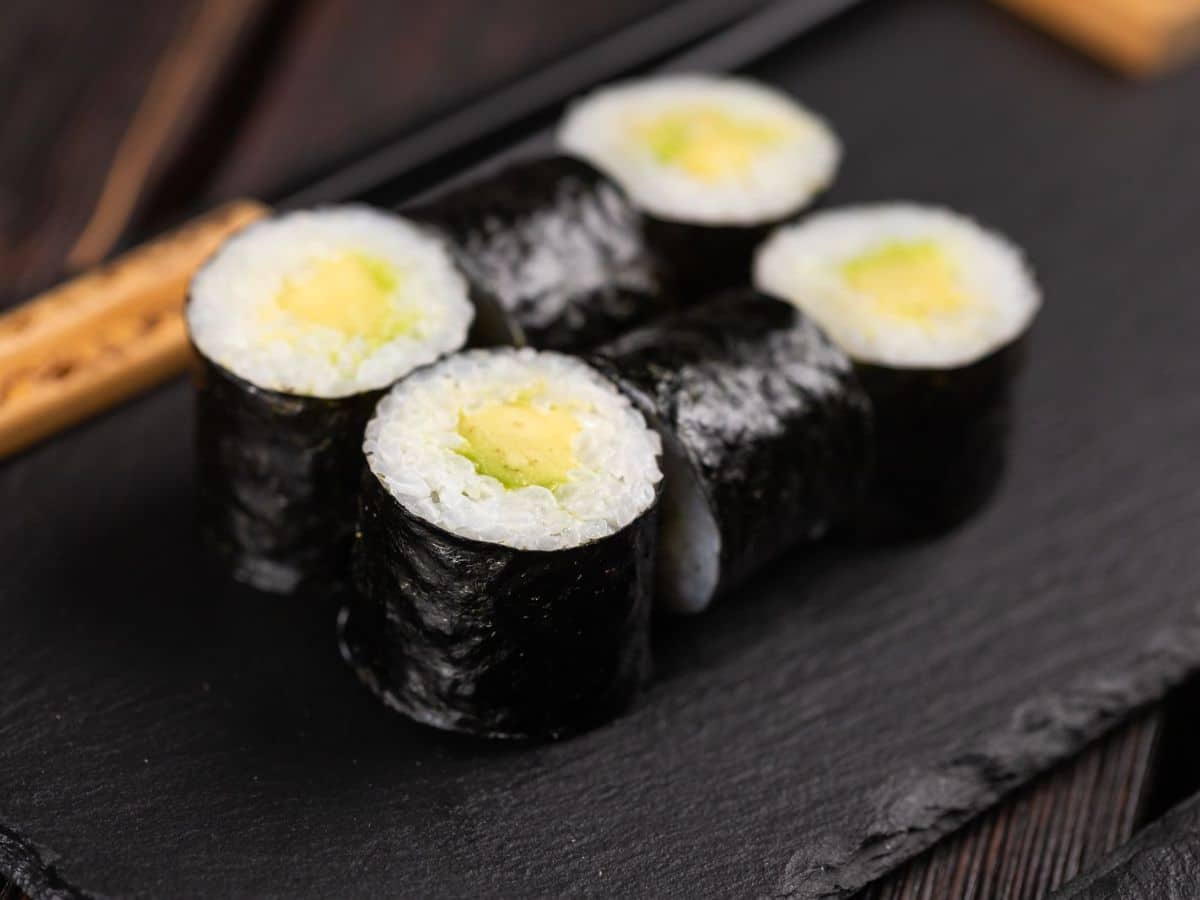 Sushi rolls with rice and avocado filling wrapped in seaweed, presented on a black slate board.