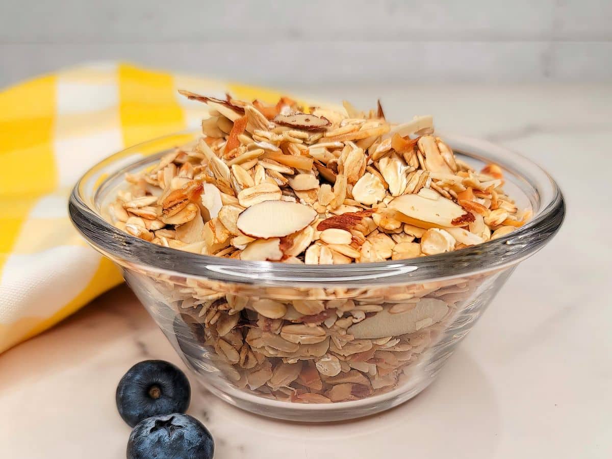 A bowl of granola with sliced almonds on a marble surface, accompanied by blueberries and a yellow-striped napkin.