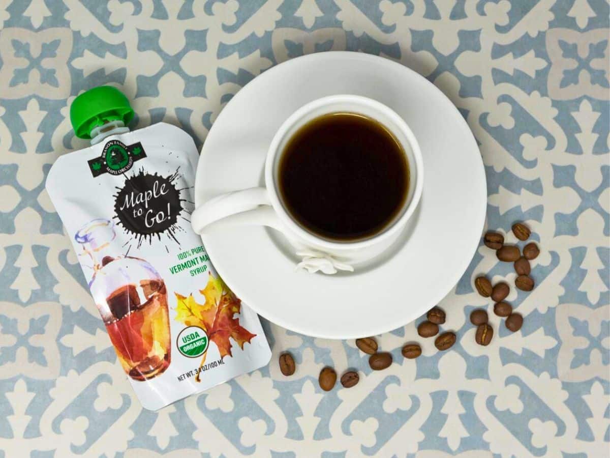 A cup of coffee on a saucer surrounded by coffee beans, next to a maple & go maple syrup pouch, on a patterned tablecloth.