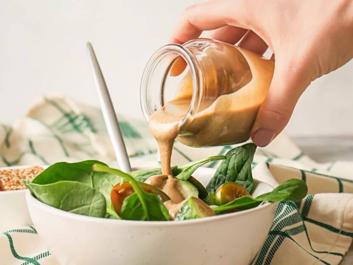 A person pours creamy dressing from a jar onto a fresh spinach and tomato salad in a white bowl.