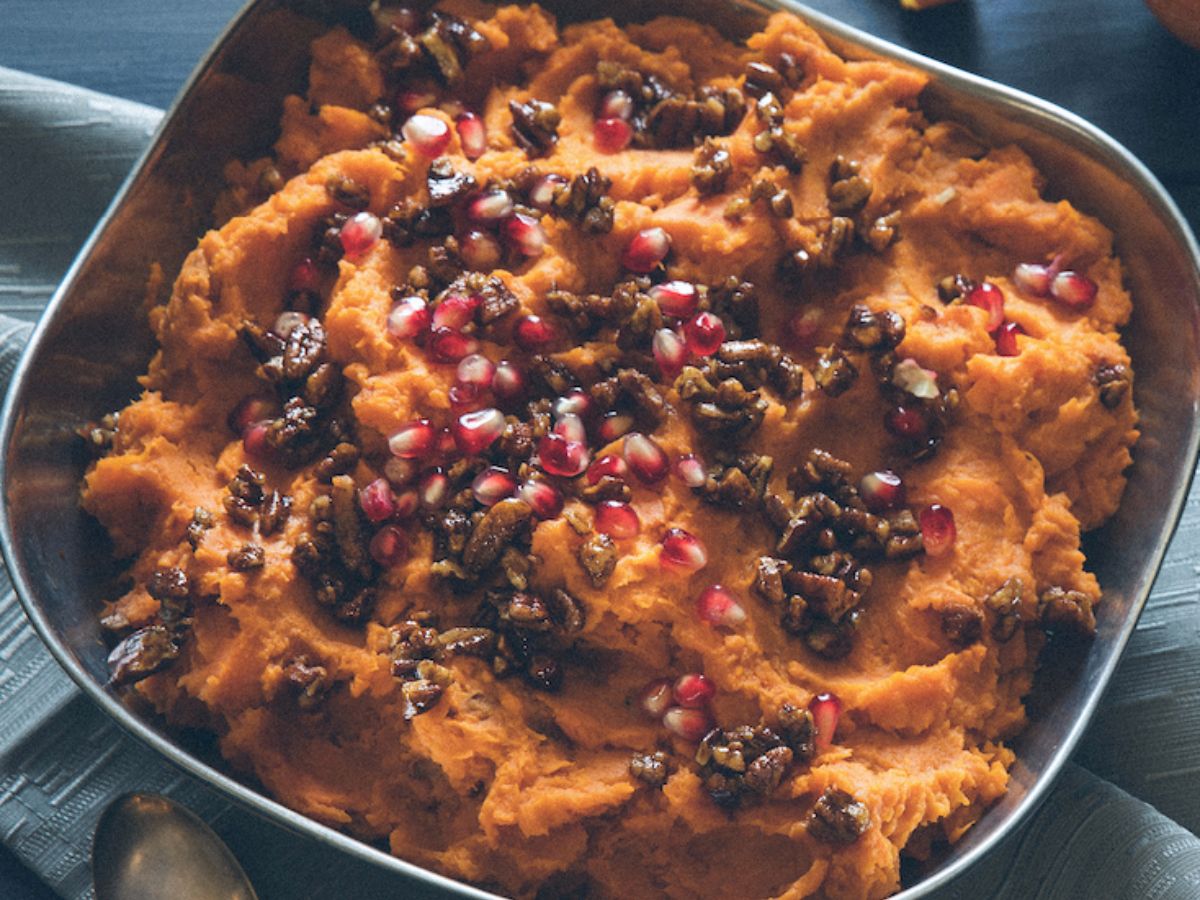 A dish of mashed sweet potatoes topped with pomegranate seeds and candied nuts, served in a rustic metal bowl with a spoon on a wooden table.