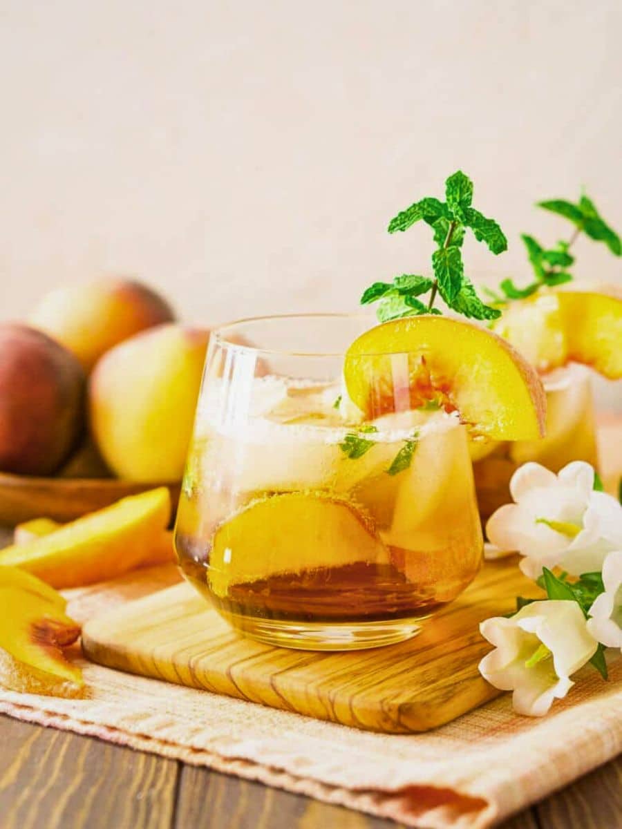 A glass of iced tea with slices of peach and a sprig of mint, surrounded by fresh peaches and white flowers on a wooden table.