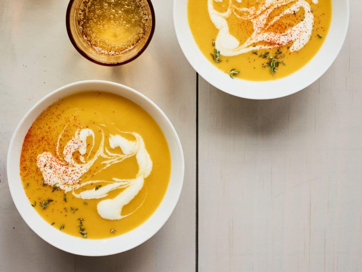 Two bowls of creamy pumpkin soup garnished with cream and spices, served with a side of honey on a wooden table.