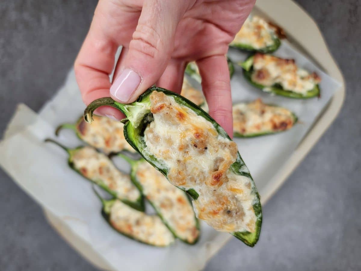 A hand holding a stuffed jalapeño pepper over a tray of similar appetizers.
