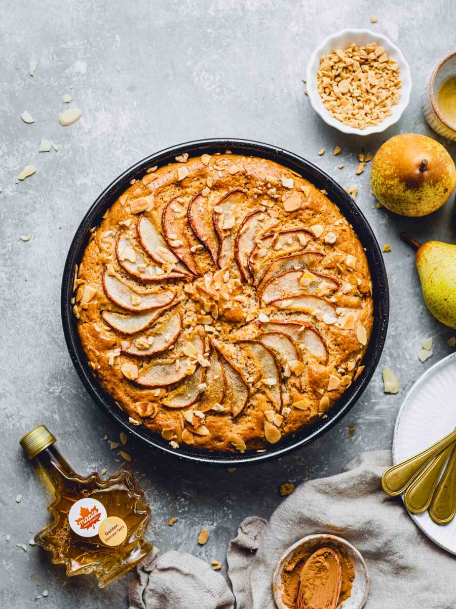 A freshly baked pear cake topped with sliced pears and almond flakes, served with honey and ground cinnamon on a gray surface.