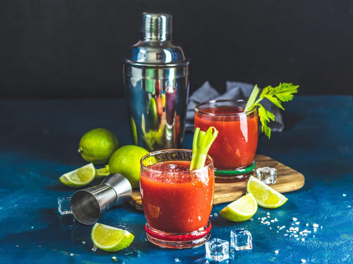 Two bloody mary cocktails on a wooden board with celery sticks, surrounded by limes, ice cubes, and a cocktail shaker on a dark blue background.