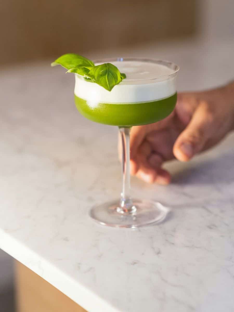 A person holding a green basil cocktail in a stemmed glass on a marble counter.
