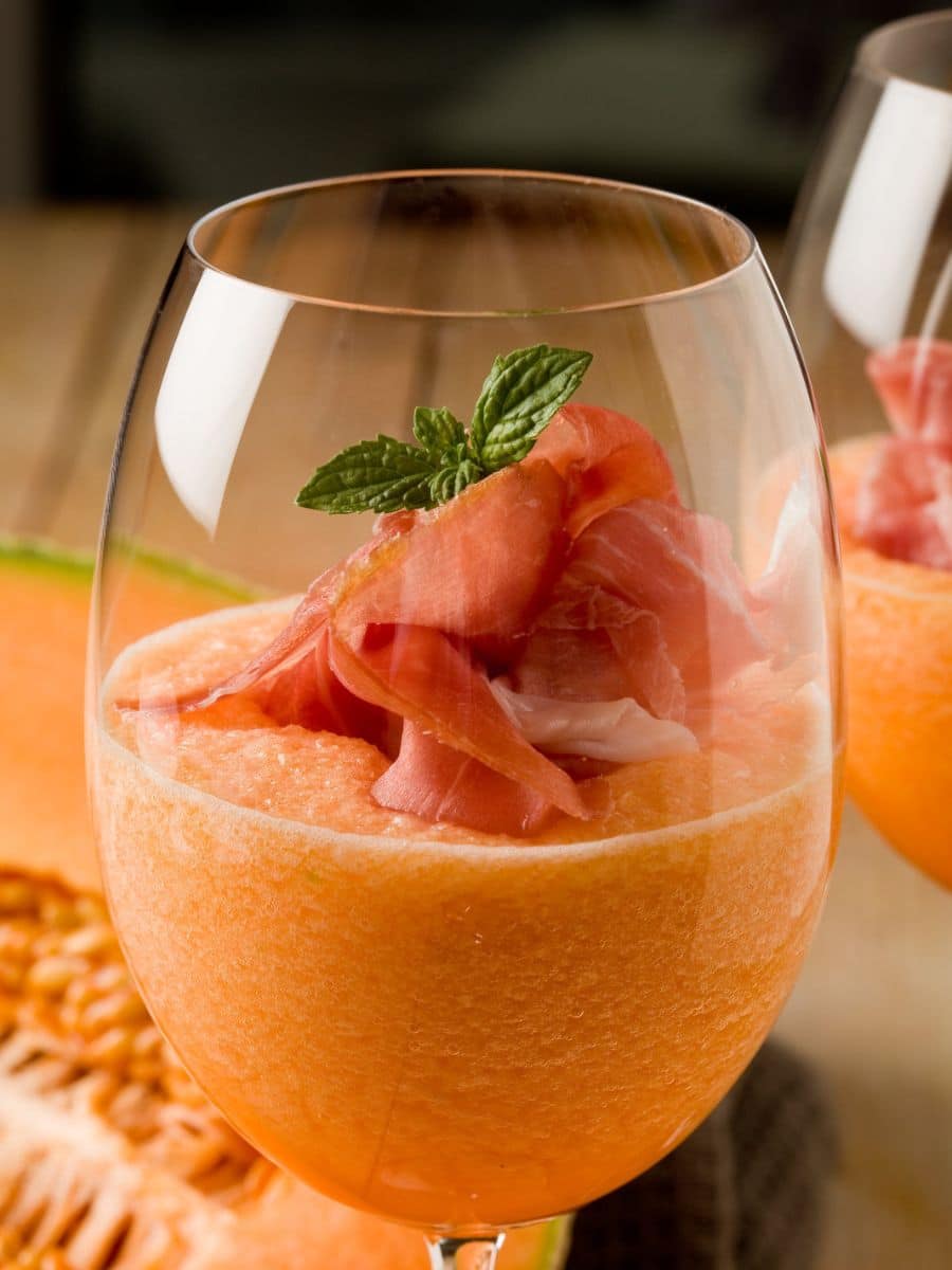 A glass of cantaloupe melon smoothie topped with prosciutto and a mint leaf, with slices of cantaloupe in the background.