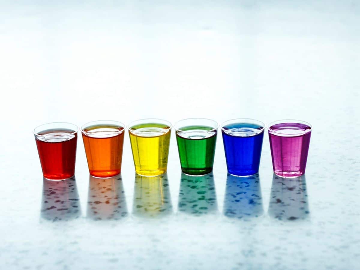 Six colorful liquids in translucent plastic cups arranged in a row on a reflective surface, displaying a spectrum from red to purple.