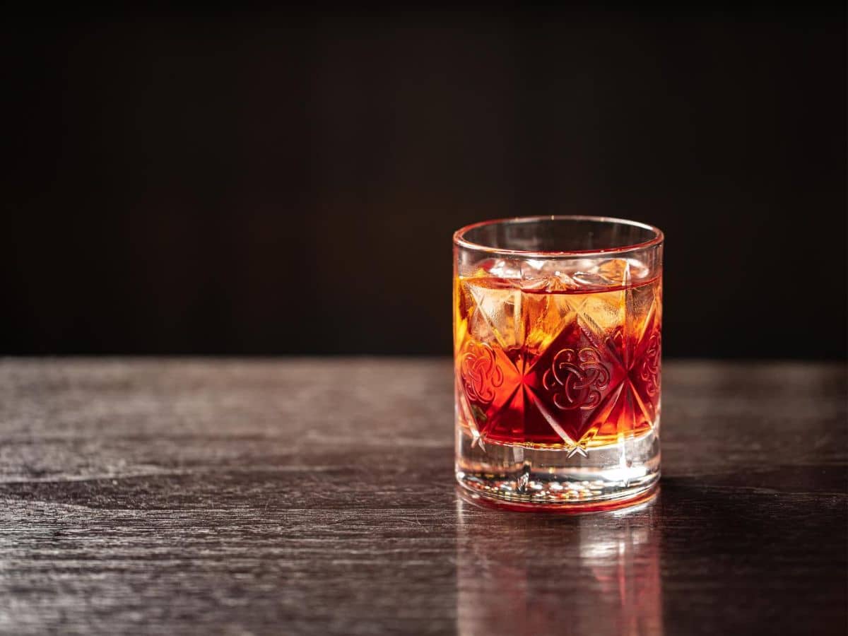 A glass of whiskey with ice on a wooden table, illuminated against a dark background.