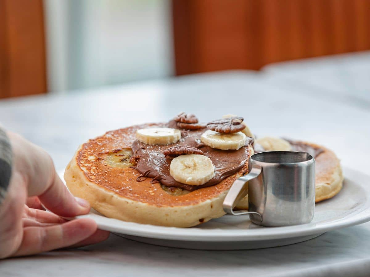 A person reaching for a plate with a pancake topped with chocolate spread, banana slices, and pecans, beside a small pitcher of syrup.