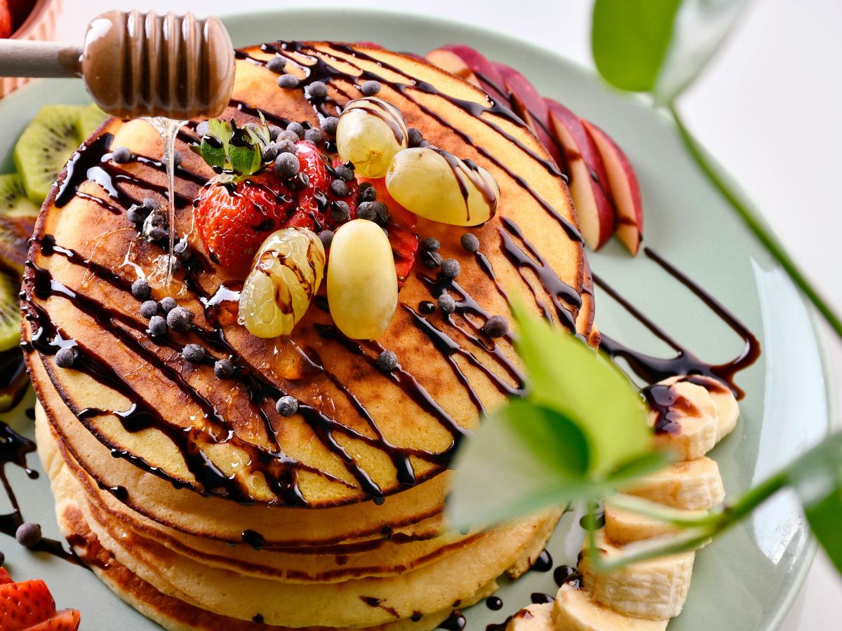A stack of pancakes topped with assorted fruits, nuts, and a drizzle of chocolate and honey, served on a green plate.