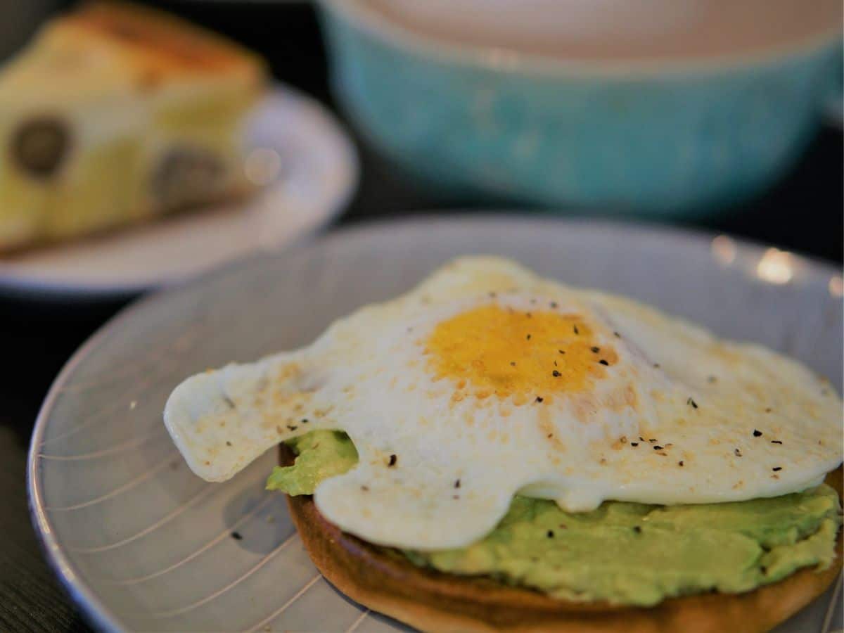 A fried egg on avocado toast, sprinkled with pepper, served on a blue-patterned plate with a slice of cake in the background.
